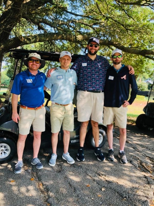 Four young men posing by golf cart under trees